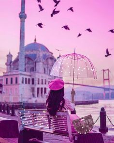 a woman sitting on a bench with an umbrella and birds flying over the water behind her