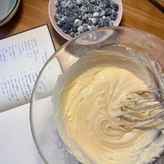 a mixing bowl filled with batter and blueberries next to an open book on a table