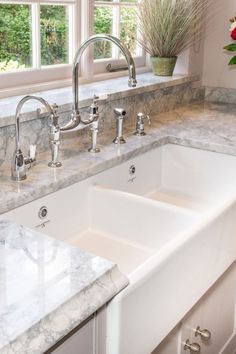 a white kitchen sink with two faucets and marble counter tops in front of a window