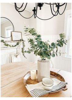 a wooden table topped with a white vase filled with greenery next to a candle