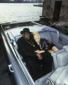 a man and woman sitting in the back of a blue convertible car next to a body of water