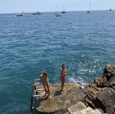 two women in bikinis standing on the edge of a cliff by the ocean with boats in the background