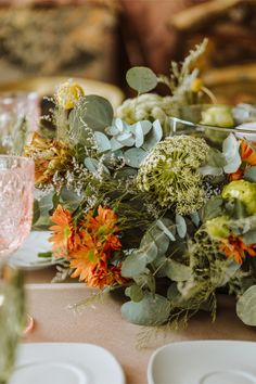 an arrangement of flowers and greenery is sitting on a table with white plates, napkins and wine glasses