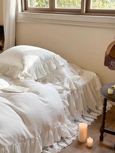 an unmade bed with white ruffled bedspread and candles on the floor