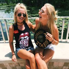 two beautiful young women sitting next to each other on a wall near water and trees