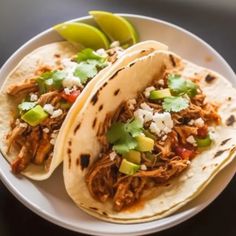 two shredded pork tacos on a white plate with limes and cilantro