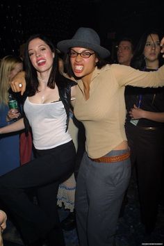 two women are dancing at a party with people in the background and one is wearing a cowboy hat