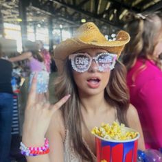 a woman in a cowboy hat and glasses holding a popcorn bucket with her fingers up