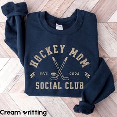 the hockey mom social club sweatshirt is laying on top of a wood floor and has two crossed sticks sticking out of it