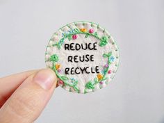 a hand holding a small embroidered badge with the words reduce reuse recycle