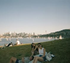 three people sitting on the grass in front of a body of water with sailboats in the background