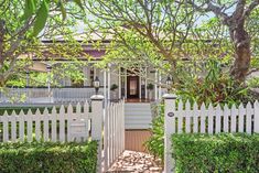 a white house with a picket fence and trees in the front yard, surrounded by greenery