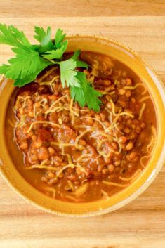 a yellow bowl filled with chili and beans on top of a wooden table next to a green leafy garnish