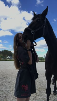 a woman standing next to a black horse on top of a sandy field with clouds in the background