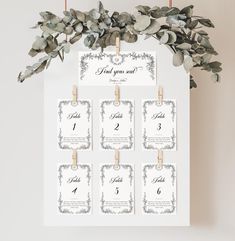 wedding seating cards with greenery and wooden pegs hanging on a white board in front of a wall