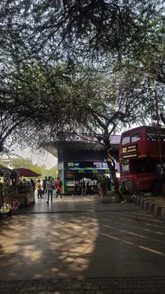 people are walking around in the shade under some trees and busses at an open air market