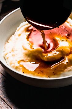 a person pouring sauce over mashed potatoes in a white bowl on a wooden table