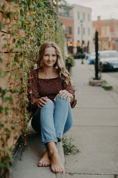 a woman sitting on the sidewalk next to a brick wall with ivy growing up it