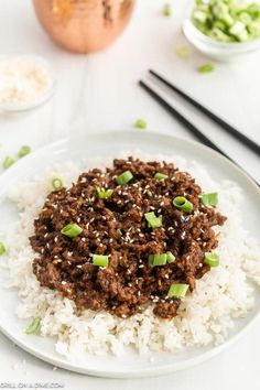 a white plate topped with rice covered in ground beef and green onions next to chopsticks