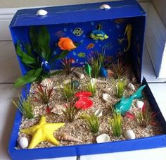 an open blue box filled with plants and fish on top of dirt covered ground next to a window