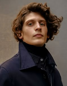 a young man with curly hair wearing a peal coat and black turtle neck sweater