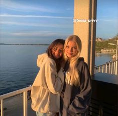 two young women standing next to each other on a balcony near the water and looking at the camera