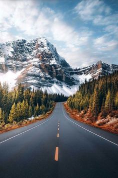 an empty road with mountains in the background and trees on both sides that are surrounded by snow