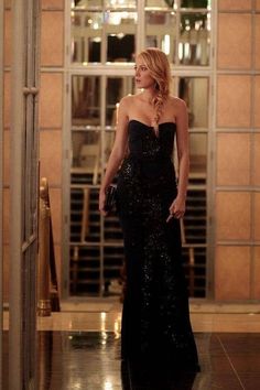 a woman in a black dress is walking down the hall with her hand on her hip