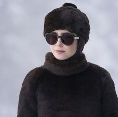 a woman wearing sunglasses and a fur hat with dark shades on her face, standing in front of a gray background