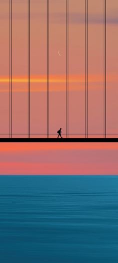 a man walking across a bridge over water at sunset with the moon in the distance