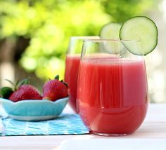 two glasses filled with red liquid next to strawberries and cucumbers