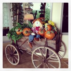 a doll sitting on top of a wooden wagon filled with pumpkins and plants in front of a window