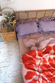 an unmade bed with purple and red sheets on it, next to a wicker night stand