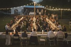 a large group of people sitting at tables in the middle of a field with lights strung over them