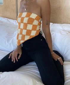 a woman sitting on top of a bed wearing black pants and an orange knitted top