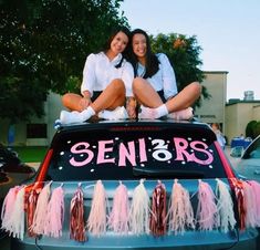 two women sitting on the back of a car with pink tassels hanging from it