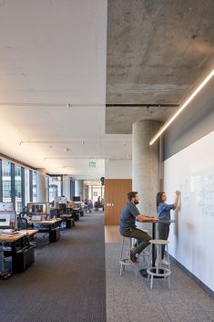 two people sitting at desks writing on a whiteboard in an open office space