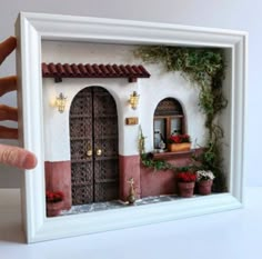 a hand is holding up a miniature house with potted plants on the windowsill