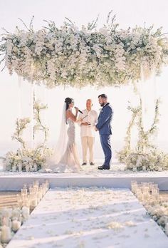 a man and woman standing under a white flower covered wedding arch