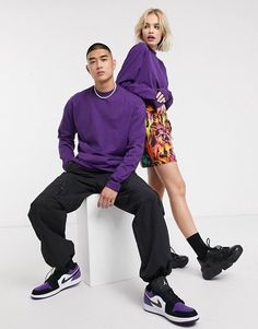 a man and woman sitting on top of a white block wearing purple sweaters, black pants and sneakers