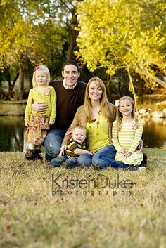 a family sitting on the grass in front of a pond with trees and water behind them