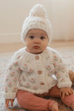 a baby sitting on the floor wearing a white knitted sweater and hat with pompom