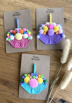 three small birthday cupcakes with candles and pom poms on the top