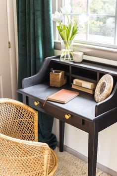 a desk with a chair, vase and flowers on it in front of a window