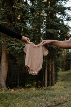 two people holding up a baby's bodysuit in the air with trees in the background