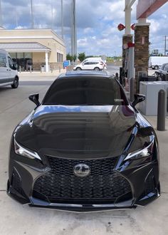 the front end of a black sports car parked at a gas station with other cars in the background