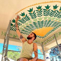 a man sitting on top of a step ladder painting a ceiling with green and yellow leaves