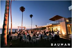 a group of people sitting at tables in front of palm trees with lights strung over them