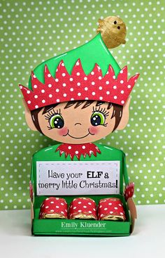 an elf doll sitting in front of a green box filled with chocolates