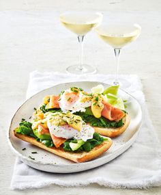 two glasses of wine are next to an open faced sandwich on a plate with salmon, avocado and lettuce